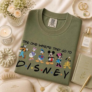 May include: Olive green t-shirt featuring Disney characters and the text "THE ONE WHERE THEY GO TO DISNEY". The shirt has a crew neck and short sleeves. The image includes accessories like jewelry, a candle, and a perfume bottle.