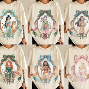 May include: Cream-colored t-shirts featuring watercolor portraits of Disney princesses within ornate frames. Each shirt showcases a different princess, surrounded by decorative bows and ribbons, creating a whimsical and artistic design.