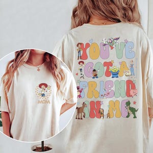Peut inclure: Un t-shirt couleur cr&egrave;me avec un petit motif circulaire de Jessie sur la poitrine et le mot MOM en dessous. Le dos pr&eacute;sente de grandes lettres color&eacute;es de style bulle formant YOU'VE GOT A FRIEND IN ME, entour&eacute;es de divers personnages Toy Story.
