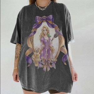 May include: Dark gray t-shirt featuring a framed illustration of a princess in a purple dress holding a frying pan, with a green chameleon. The frame has purple bows and ribbons. The shirt is a casual, oversized style.