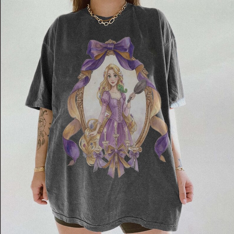 May include: Dark gray t-shirt featuring a framed illustration of a princess in a purple dress holding a frying pan, with a green chameleon. The frame has purple bows and ribbons. The shirt is a casual, oversized style.