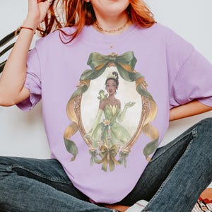May include: A lilac-colored t-shirt featuring a framed illustration of a princess in a green gown, with a frog on her shoulder. The frame is adorned with a green bow and gold ribbons. The princess is wearing a crown and gloves.