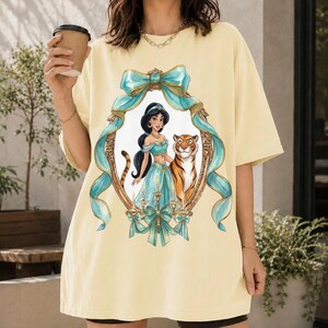 May include: A light yellow t-shirt featuring a framed illustration of Princess Jasmine and a tiger. The frame is adorned with teal ribbons and gold accents. The princess is wearing a teal outfit. The shirt has a relaxed fit.