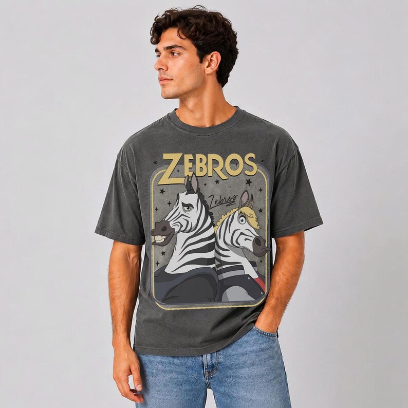May include: A charcoal gray short-sleeve t-shirt featuring a graphic print of two cartoon zebras. The text "ZEBROS" appears in large yellow letters above the zebras, with a smaller "Zebros" script below. The design is enclosed in a rounded rectangle.