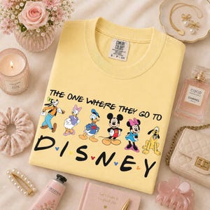 May include: A yellow Comfort Colors t-shirt featuring Disney characters and the text "THE ONE WHERE THEY GO TO DISNEY." The design includes Goofy, Daisy Duck, Donald Duck, Mickey Mouse, Minnie Mouse, and Pluto. The shirt is surrounded by accessories.