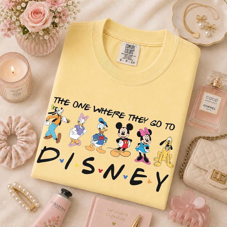 May include: A yellow Comfort Colors t-shirt featuring Disney characters and the text "THE ONE WHERE THEY GO TO DISNEY." The design includes Goofy, Daisy Duck, Donald Duck, Mickey Mouse, Minnie Mouse, and Pluto. The shirt is surrounded by accessories.