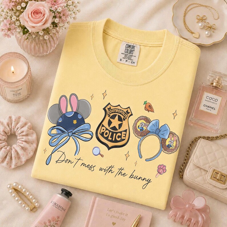 May include: A pale yellow t-shirt featuring a graphic print of a Zootropolis police badge, two pairs of mouse ears, and the text "Don't mess with the bunny." The shirt has a Comfort Colors label at the inner neckline and is displayed on a flat surface.