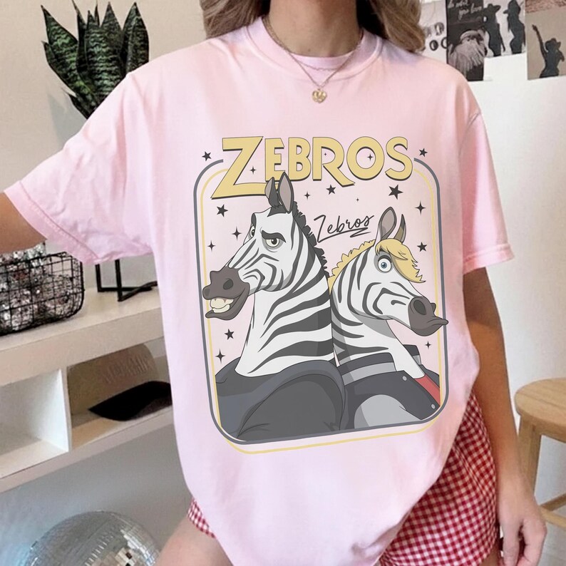May include: A light pink short-sleeve t-shirt featuring a graphic print of two cartoon zebras inside a rectangular frame. The text "ZEBROS" appears in large yellow letters above the zebras, with "Zebros" in script below. The zebras have black and white stripes.