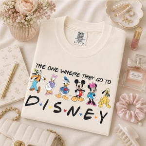 May include: Cream-colored sweatshirt featuring the text "THE ONE WHERE THEY GO TO DISNEY" with cartoon characters. The design includes Goofy, Daisy Duck, Donald Duck, Mickey Mouse, Minnie Mouse, and Pluto. The sweatshirt has a Comfort Colors label.
