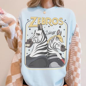 May include: A light blue crewneck sweatshirt featuring a graphic print of two cartoon zebras standing back-to-back. The text "ZEBROS" appears in large yellow letters above the zebras, with the word "Zebros" in script below, surrounded by stars.