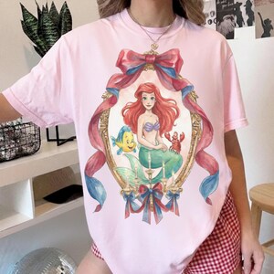 May include: A light pink t-shirt featuring a framed illustration of Ariel from The Little Mermaid. The illustration includes Ariel, Flounder, and Sebastian, with a red and blue ribbon design. The shirt is a casual style.