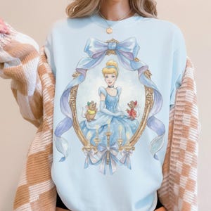 May include: Light blue sweatshirt featuring a framed illustration of Cinderella in a blue gown, surrounded by mice and candles. The frame is gold with blue ribbons and bows. The sweatshirt has a crew neck.