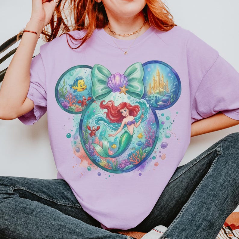 May include: A light purple t-shirt featuring a large graphic of a mermaid with red hair and a green tail, surrounded by underwater elements like coral, fish, and a castle, all arranged in a Mickey Mouse ear shape with a large teal bow.