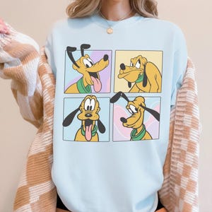 Puede incluir: Una sudadera de cuello redondo en color azul claro con un estampado gr&aacute;fico de cuatro paneles distintos, cada uno mostrando una expresi&oacute;n facial diferente del perro de dibujos animados amarillo Pluto, que lleva un collar verde. La sudadera se lleva bajo una rebeca beige y blanca.