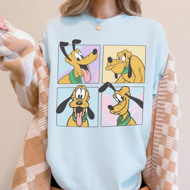 Puede incluir: Una sudadera de cuello redondo en color azul claro con un estampado gr&aacute;fico de cuatro paneles distintos, cada uno mostrando una expresi&oacute;n facial diferente del perro de dibujos animados amarillo Pluto, que lleva un collar verde. La sudadera se lleva bajo una rebeca beige y blanca.