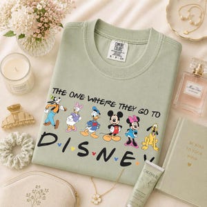 May include: Sage green crew neck t-shirt with the text "THE ONE WHERE THEY GO TO DISNEY" and Disney characters. The shirt is surrounded by accessories including a candle, perfume, and a book.