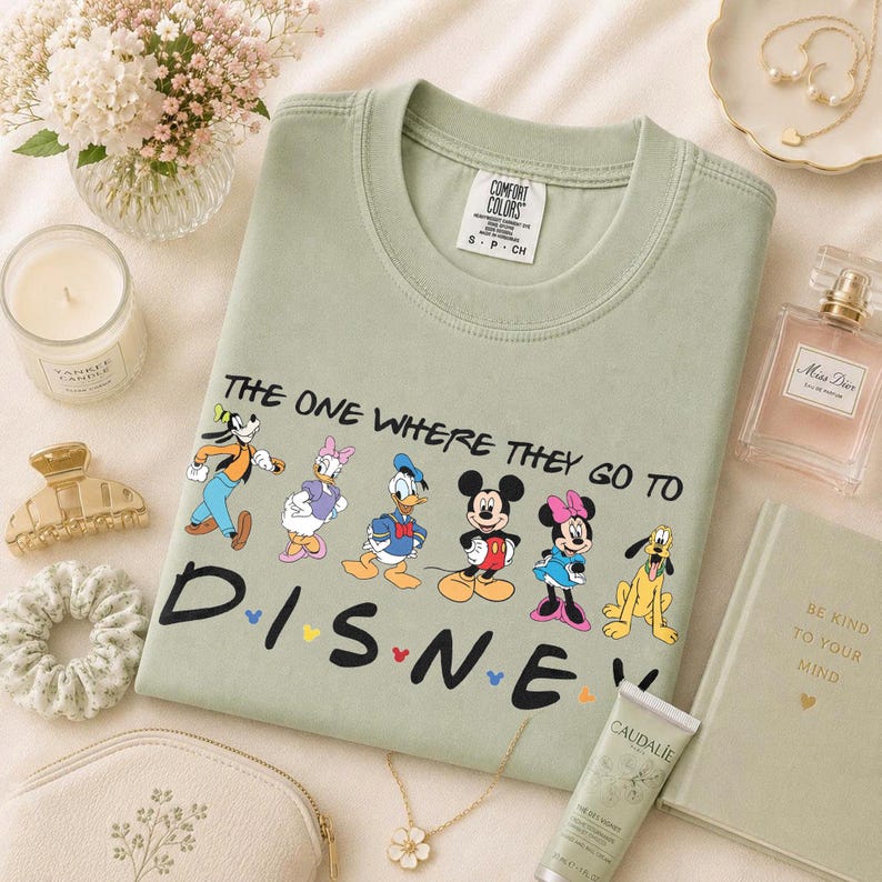 May include: Sage green crew neck t-shirt with the text "THE ONE WHERE THEY GO TO DISNEY" and Disney characters. The shirt is surrounded by accessories including a candle, perfume, and a book.