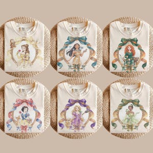 May include: Six cream-colored t-shirts featuring watercolor illustrations of Disney princesses. Each shirt displays a different princess within a decorative frame with ribbons and bows. The princesses include Belle, Pocahontas, Merida, Snow White, Rapunzel, and Tiana.