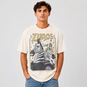 May include: An off-white short-sleeve t-shirt featuring a graphic print of two cartoon zebras. The text "ZEBROS" appears in large gold letters above the zebras, with the word "Zebros" in script below. The design is enclosed in a rectangular frame.