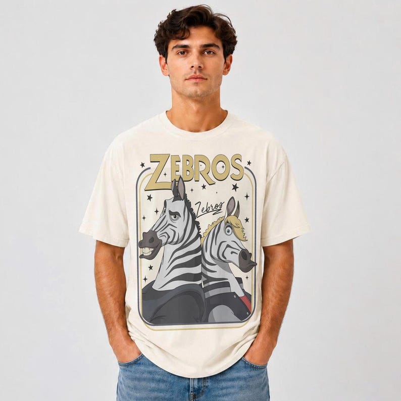 May include: An off-white short-sleeve t-shirt featuring a graphic print of two cartoon zebras. The text "ZEBROS" appears in large gold letters above the zebras, with the word "Zebros" in script below. The design is enclosed in a rectangular frame.