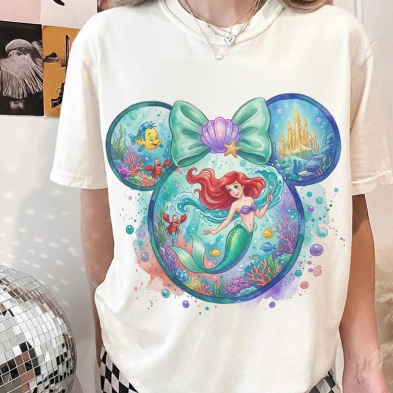 May include: A cream-colored short-sleeve t-shirt featuring a large graphic of a mermaid with red hair and a green tail, surrounded by underwater elements including a yellow fish, a red crab, a castle, and a large teal bow with a purple shell.