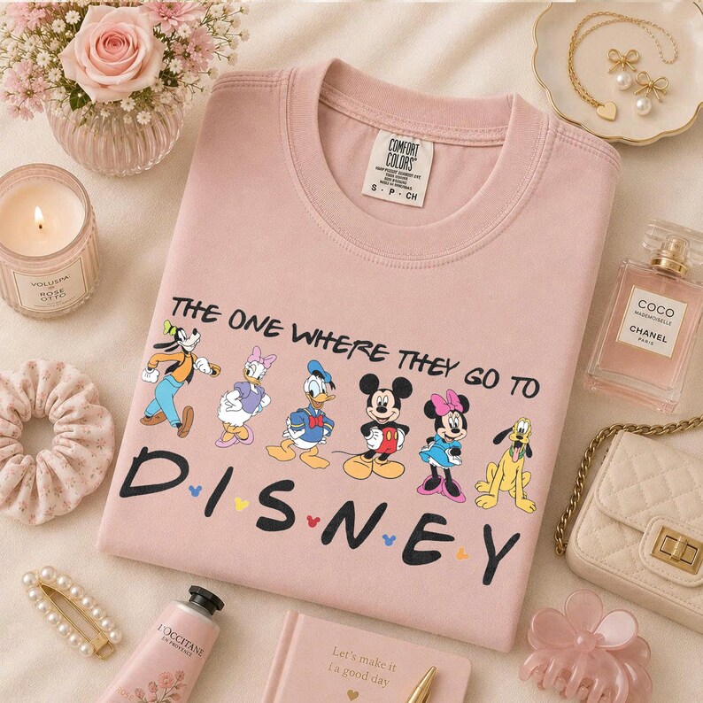 May include: Pink Comfort Colors sweatshirt featuring Disney characters and the text "THE ONE WHERE THEY GO TO DISNEY". The design includes Goofy, Daisy Duck, Donald Duck, Mickey Mouse, Minnie Mouse, and Pluto. The sweatshirt is surrounded by accessories.