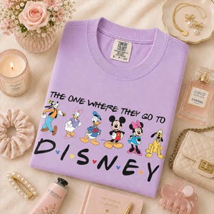 May include: A lilac Comfort Colors t-shirt featuring Disney characters and the text "THE ONE WHERE THEY GO TO DISNEY". The shirt has a crew neck and short sleeves. The design includes Mickey Mouse, Minnie Mouse, Donald Duck, and Pluto.