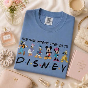 May include: A light blue t-shirt featuring the text "THE ONE WHERE THEY GO TO DISNEY" with cartoon characters. The shirt has a crew neck and short sleeves. The characters include Goofy, Daisy Duck, Donald Duck, Mickey Mouse, Minnie Mouse, and Pluto.