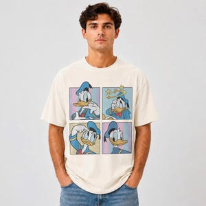 Puede incluir: Una camiseta de manga corta color crema con un estampado gr&aacute;fico de Donald Duck en cuatro paneles distintos. Cada cuadrado muestra al personaje en una pose y expresi&oacute;n diferente, incluyendo una con estrellas alrededor.