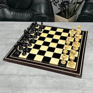 Puede incluir: Un juego de ajedrez de madera con casillas negras y crema. Las piezas de ajedrez son negras y crema, dispuestas sobre el tablero. El tablero tiene un borde marrón oscuro con un acento crema, de unos 40 cm x 40 cm.