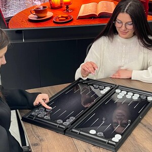 May include: A black backgammon board with a carbon fiber texture, featuring white and black triangular points. The game set includes translucent clear and opaque white circular checkers, along with two small black dice positioned on the board surface.