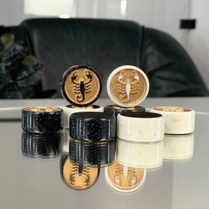 May include: A collection of decorative bottle stoppers. The stoppers feature a gold scorpion design on a black or white background. The stoppers are cylindrical and have a gold rim. The stoppers are displayed on a reflective surface.