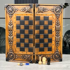 May include: A wooden chess and backgammon board set. The board is hinged and features a detailed carved floral and lion design. The chess board has black and brown squares. Chess pieces and dice are visible at the bottom.