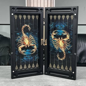Puede incluir: Tablero de backgammon con marco negro y diseño detallado de escorpión. El escorpión es de color dorado, sobre un fondo azul y negro. El tablero está abierto, revelando la superficie de juego con elementos decorativos.