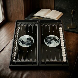 以下が含まれることがあります： ゲーム盤面に月のデザインが施された黒いバックギャモンセット。銀色のゲームピースと装飾的な銀色のアクセントが含まれています。開いた本、ペン、燭台が背景にあります。