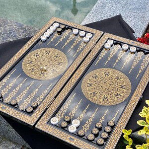 May include: A backgammon set with a black playing surface and gold decorative trim. The board features a central gold mandala design and gold accents. White and black game pieces are arranged on the board, along with a pair of dice.
