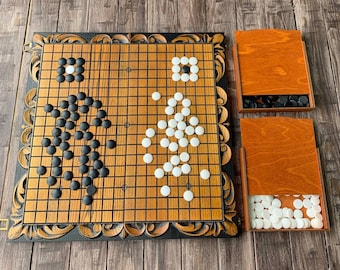 May include: A wooden game board with black and white playing pieces. The board is decorated with an ornate, carved frame. The board is set up for a game of Go.