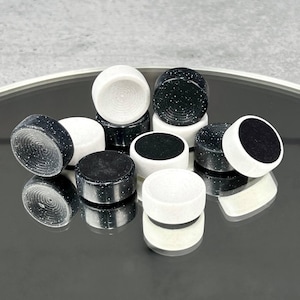 May include: A collection of black and white game pieces, possibly for checkers or backgammon, are arranged on a reflective surface. The pieces are cylindrical, with some featuring a speckled pattern and others a solid color. The pieces are approximately 1 inch in diameter.