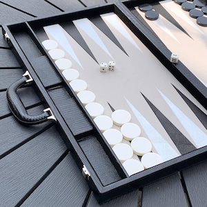Puede incluir: Un juego de backgammon negro con fichas blancas y negras. El tablero tiene un fondo gris claro con patrones triangulares negros y blancos. Hay dos dados en el tablero y el juego incluye un asa y compartimentos de almacenamiento.
