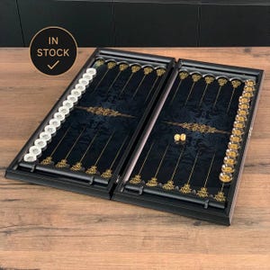 May include: An open backgammon board game set featuring a black case with a dark blue interior, gold filigree patterns, and thin lines. The set includes clear and gold-toned circular checkers arranged along the sides and two small dice in the center. A circular badge in the top left corner displays the text IN STOCK with a checkmark.