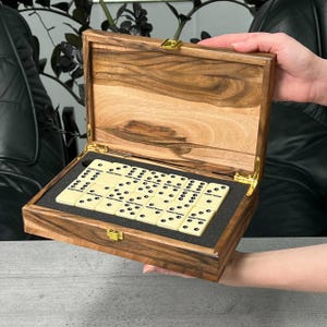 Peut inclure: Un jeu de dominos en bois dans une boîte décorative. La boîte est en bois brun clair avec un motif de grain naturel et des charnières et un fermoir dorés. Les dominos sont de couleur ivoire avec des points noirs.