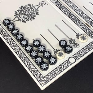 Puede incluir: Un tablero de backgammon con piezas en blanco y negro. El tablero es blanco con un borde decorativo negro y elementos de diseño. Las piezas del juego son negras con diseños de estrellas blancas. El tablero está parcialmente abierto.