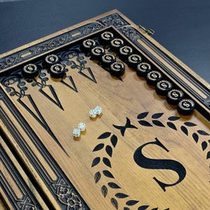 May include: A wooden backgammon board with black and gold accents. The board has a carved design and features a wreath with the letter "S" in the center. There are white dice on the board.