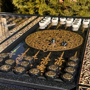 Puede incluir: Primer plano de un tablero de backgammon con detalles en negro y dorado. El tablero presenta patrones decorativos dorados, fichas blancas y negras, y dos dados. El juego está configurado y listo para jugar.