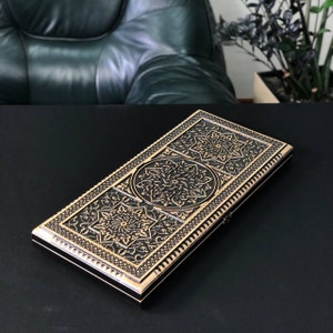 May include: A black wooden box with intricate gold-colored designs. The box has a rectangular shape and a raised border. The designs are floral and geometric.