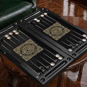 May include: A black backgammon board featuring gold-colored triangular points and circular game pieces in black and white. The center of the board displays a gold graphic emblem with the text "VINTAGE STYLE ART DECO FRAME DESIGN" enclosed in a decorative border.