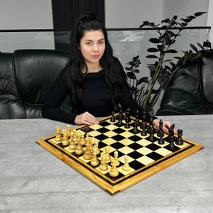 Puede incluir: Un juego de ajedrez de madera con casillas blancas y negras y piezas de ajedrez. El tablero tiene un borde de madera clara con una incrustación de madera oscura. Las piezas de ajedrez son blancas y negras. El juego está sobre una superficie gris.