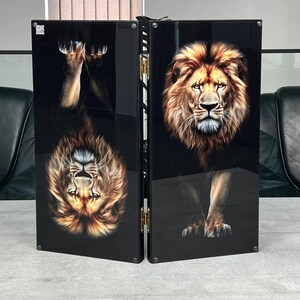 May include: A folding game board featuring a high-gloss black finish with a detailed portrait of a lion on the right panel and an inverted reflection of the same lion on the left panel. The two panels are connected by gold-coloured metal hinges.