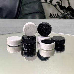 May include: A set of black and white checkers with a speckled finish. The checkers are arranged in a pyramid shape on a reflective surface.