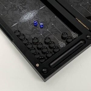 May include: A black backgammon board with a decorative floral pattern in silver and gray. The board includes black game pieces, two blue dice, and a wooden dice cup. The board is set against a white background.